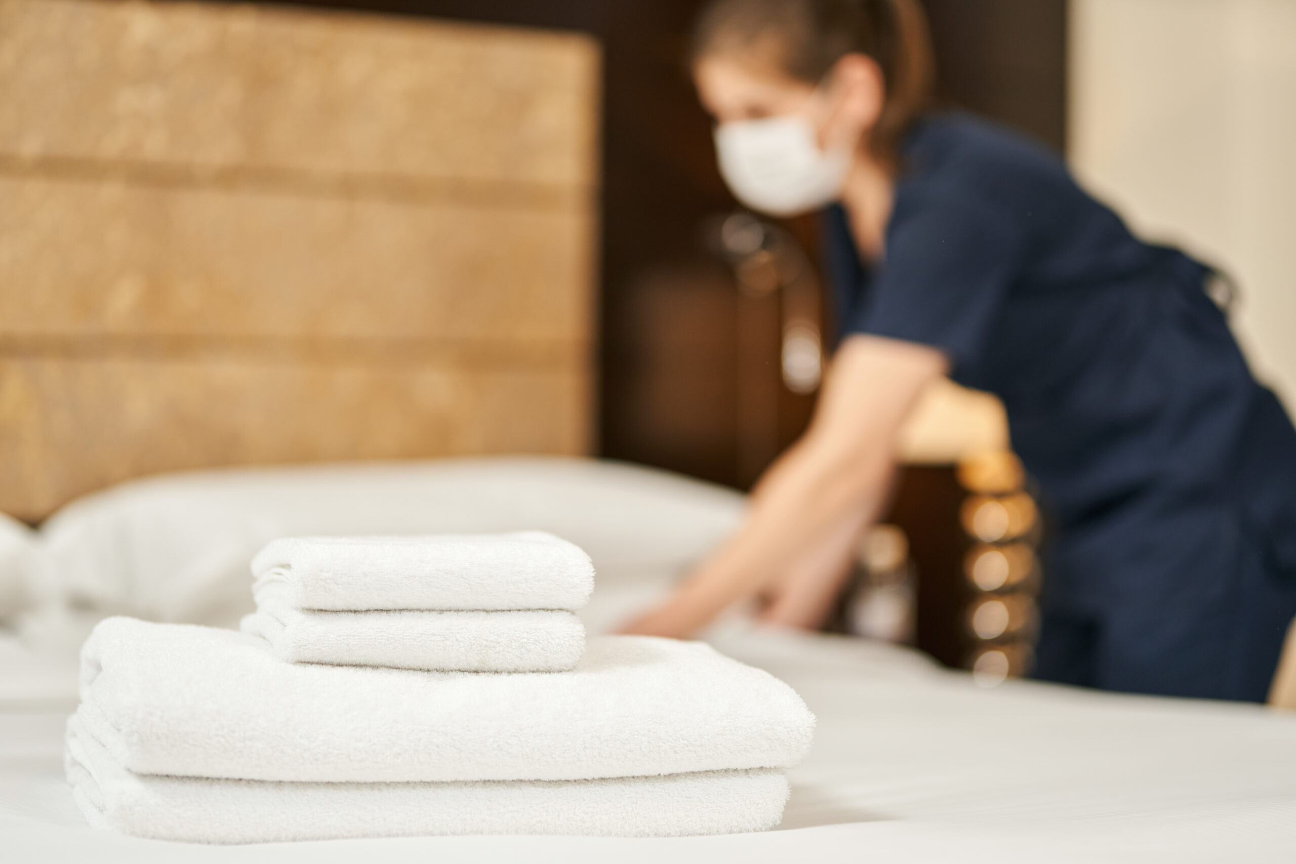 White fresh towels on the bed with a maid in a mask on the background. Hotel service concept.